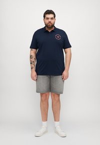 Tricou polo bleumarin cu mâneci scurte și detaliu logo rotund; purtat cu pantaloni scurți gri și teniși albi, stând în fața unui fundal simplu.