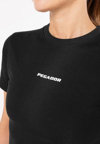 Čierne bavlnené tričko s okrúhlym výstrihom a krátkymi rukávmi. Obsahuje logo "PEGADOR" v bielej farbe, umiestnené mierne diagonálne cez hruď.