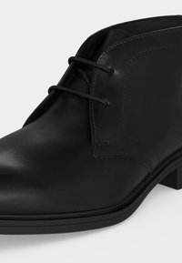 Chaussure habillée pour homme en cuir noir avec bout arrondi, couture minimale et deux œillets pour lacets noirs, présentée sur fond blanc.
