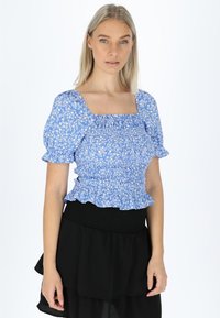 Blå blomstret bluse med pufærmer, firkantet halsudskæring og smocket talje. Har en flæse i bunden. Bæres med en sort lagdelt nederdel.