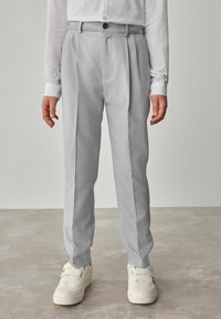 Pantaloni grigi chiari in tessuto testurizzato, con pieghe e chiusura a bottone, abbinati a sneakers bianche. Design a gamba dritta.