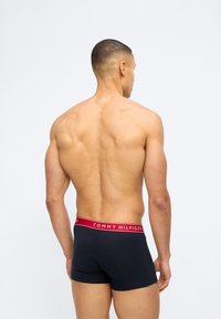 Μυώδης άνδρας, φαίνεται από πίσω, φοράει μαύρα μποξεράκια Tommy Hilfiger με κόκκινη ζώνη μέσης, στέκεται μπροστά σε απλό λευκό φόντο.