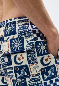 Κοντινό πλάνο ενός χεριού που ακουμπά κοντά στην πίσω τσέπη ενός μοτίβο σορτς με σχέδια από ήλιους, φεγγάρια, αστέρια, ψάρια και κύματα σε αποχρώσεις μπλε, λευκού και καφέ.