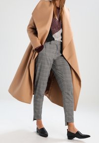 Beige wikkeljas over een bordeauxrode en grijze gebreide trui, gecombineerd met grijze geruite broek en zwarte instappers. Soepele texturen en een op maat gemaakte pasvorm.