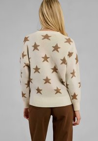 Maglione crema dalla vestibilità morbida, con motivi di stelle bronzee in tutto il capo. I polsini e l'orlo a coste aggiungono texture. Indossato con pantaloni marroni.