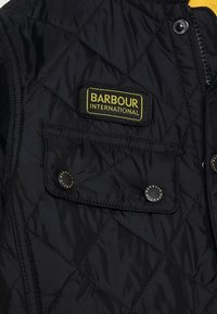 Schwarze gesteppte Jacke mit einem gelben und schwarzen "Barbour International"-Logo. Verfügt über einen oberen Reißverschluss und eine Fronttasche mit Druckknopfverschluss.