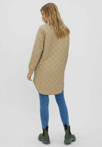 Veste longue matelassée beige avec des poignets côtelés, dotée d'un ourlet arrondi ; portée sur un jean bleu et des bottines vertes.