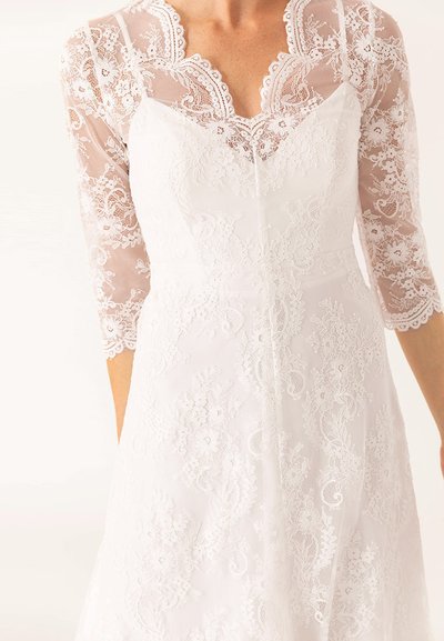 Femeie îmbrăcată într-o rochie de mireasă din dantelă albă, cu mâneci transparente până la trei sferturi și broderie florală pe bodice și fustă.