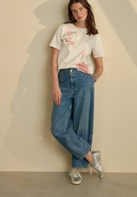 T-shirt en coton blanc avec une broderie florale colorée, associé à un jean en denim bleu taille haute et ample, et des baskets gris clair.