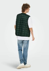 Gilet rembourré matelassé vert avec un motif en carrés, porté sur un t-shirt blanc et associé à un jean bleu clair et des sneakers beiges. Vue de dos.