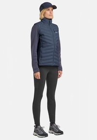 Marineblauw puffer-vest met verticale doorgestikte patronen, gedragen over een top met lange mouwen en gecombineerd met zwarte leggings en gestructureerde wandelschoenen.