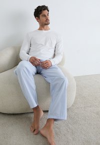 Cămașă albă cu mâneci lungi, asortată cu pantaloni de pijama cu dungi albastre și albe, așezat pe un scaun rotund texturat de culoare crem.