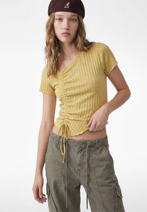 Jonge vrouw met golvend haar, gekleed in een mosterdkleurige, geplooide top met korte mouwen, kaki cargobroek en een donkerbruine Kangol buckethoed tegen een witte achtergrond.