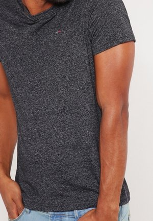 Mand iført en tætsiddende mørkegrå t-shirt med korte ærmer og et lille rødt, hvidt og blåt logo på brystet, kombineret med lyseblå jeans.