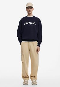 Sudadera navy con letras blancas "DESIGUAL", complementada con pantalones cargo beige con bolsillos laterales y zapatillas crema.