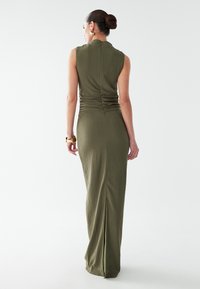 Robe maxi sans manches vert olive en tissu texturé, avec des fronces au dos et une fente latérale pour faciliter les mouvements.