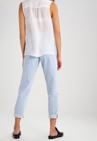 White sleeveless blouse with a back yoke, paired with light blue rolled-up trousers and black slip-on shoes. Smooth, lightweight fabric.