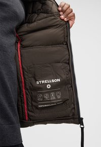Giacca imbottita marrone con design trapuntato. Presenta una fodera rossa e una tasca frontale. L'etichetta mostra il marchio "STRELLSON" e le informazioni sul materiale.