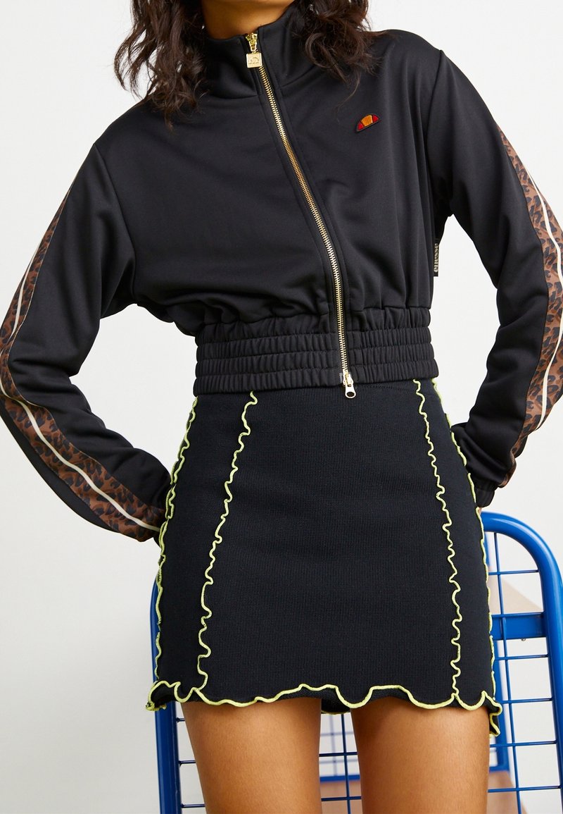 Femme portant une veste zippée noire courte avec des manches imprimées léopard et une jupe noire à bordure ondulée jaune, debout les mains sur les hanches.