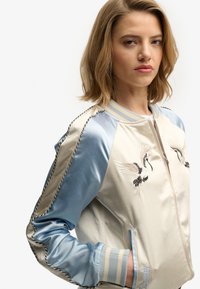 Bomber in raso color crema con maniche in raso azzurro chiaro. Presenta gru ricamate, polsini a righe e chiusura con zip. Texture liscia.