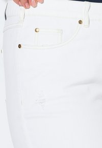 Jeans de mezclilla blanca que presentan un área desgastada, remaches de latón y costuras en contraste. Diseñados con un estilo clásico de cinco bolsillos.