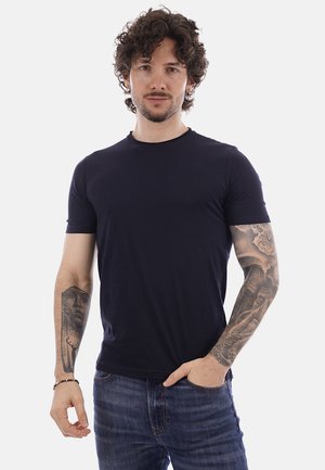 Uomo con capelli ricci e orecchini, indossa una t-shirt nera e jeans, mostra tatuaggi dettagliati su entrambe le braccia, in piedi davanti a uno sfondo bianco.