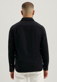 Man met kort haar, gekleed in een zwart shirt met lange mouwen en een witte broek, gezien van achteren tegen een effen lichte achtergrond.