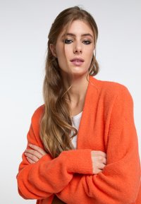 Överdimensionerad orange cardigan med mjuk textur, ribbade ärmslut och djup V-ringning. Burit över en enkel vit skjorta, med armarna i kors.