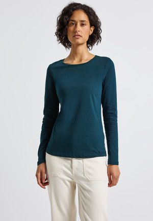 Donna con capelli ricci scuri che indossa una maglietta a maniche lunghe color teal e pantaloni bianco sporco, in piedi davanti a uno sfondo bianco semplice.