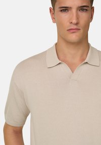 Jeune homme portant un polo beige à manches courtes avec un col en V, faisant face à l'avant sur un fond neutre.