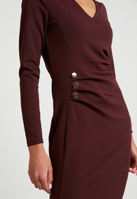 Burgundy klänning med V-ringning, långa ärmar och sidoforryning. Har tre metallknappar längs den samlade sömmen vid höften.