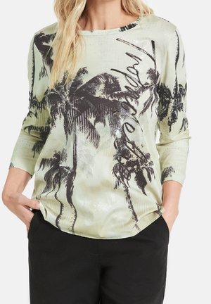 Femme portant une chemise à manches longues vert clair avec un imprimé de palmier noir et le mot "Beverly" en écriture cursive sur le devant, assortie à un pantalon noir.
