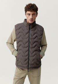 Bruine doorgestikte vest met een hoge kraag en ritssluiting, voorzien van een chevronpatroon. Gedragen over een licht, langslavend top en beige broek.