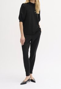 Schwarzes Kurzarmtop mit gepufften Ärmeln und hohem Ausschnitt, kombiniert mit schwarzen Slim-Fit-Jogginghosen und spitzen schwarzen Absätzen.