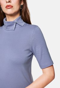 Short-sleeve, light purple top with a high collar featuring a decorative silver accent and smooth, stretchy fabric.