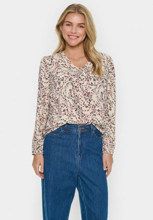 Blusa color crema con un motivo floreale vorticoso in rosso e blu navy, con scollatura a V e maniche lunghe, abbinata a jeans blu a vita alta.