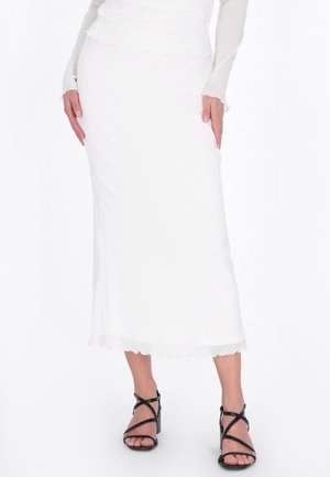 Frau trägt einen langen weißen Rock und ein durchsichtiges weißes Oberteil mit langen Ärmeln, kombiniert mit schwarzen, verstellbaren High-Heel-Sandalen.