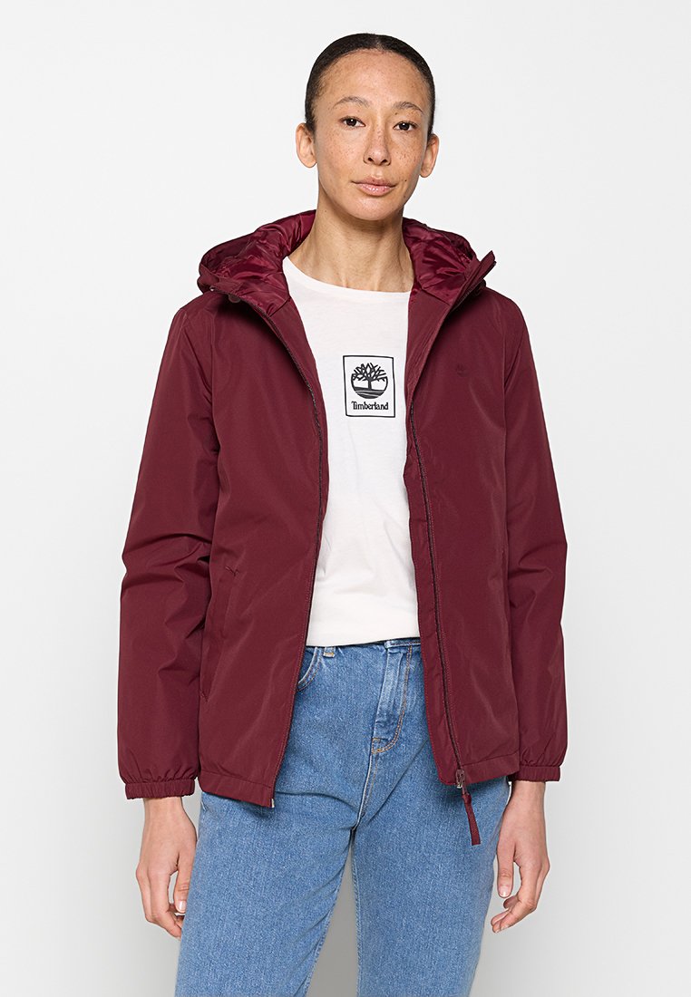 Chaqueta impermeable burdeos con capucha, que presenta puños elásticos, cierre con cremallera y una textura suave, llevada sobre una camiseta blanca y jeans.