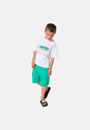 Jonge jongen die een wit T-shirt met grafisch ontwerp draagt, tealkleurige cargoshorts en zwarte slidesandalen, staand tegen een effen witte achtergrond.