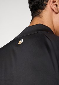 Μαύρο αθλητικό πουκάμισο με υφή υφάσματος, κοφτερό γιακά και μικρό, αντίθετο λογότ�υπο στην πλάτη κοντά στον λαιμό.