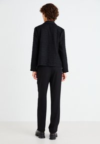 Blazer negro texturizado con solapa de pico, combinado con pantalones negros entallados y zapatos gruesos negros. Tejido cálido y tejido, silueta recta.