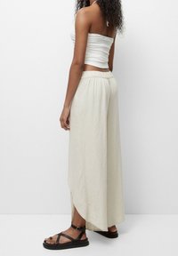 Pantalons larges de couleur beige clair en tissu doux avec une taille élastique. Comprend des fentes sur les côtés pour un meilleur confort de mouvement. Portés avec des sandales noires.
