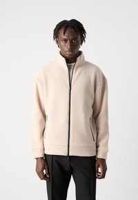 Fleece jacket in light beige, featuring a high collar, side zip pockets, and black zipper accents. Soft texture and relaxed fit.