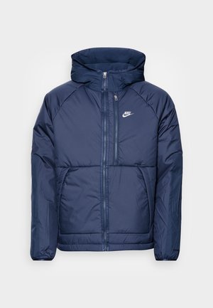 Temno modra puffer jakna z sprednjim zadrgo, visokim ovratnikom in kapuco. Ima raglan rokave in bel Nike logotip na prsih.