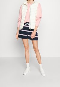 Witte gewatteerde vest, roze sweatshirt met lange mouwen, marineblauwe gestreepte shorts en witte sneakers van textielstof met een ontspannende pasvorm.