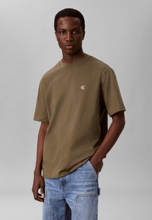 Junger Mann, der ein olivgrünes CK-T-Shirt und hellblaue Jeans trägt, steht mit einer Hand in der Tasche vor einem einfarbigen grauen Hintergrund.