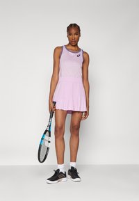 Conjunto de tenis morado con una falda plisada y una blusa texturizada. Zapatillas deportivas negras y una raqueta de tenis en la mano.