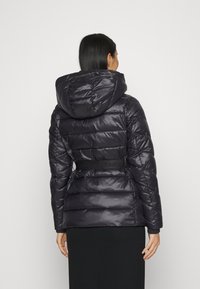 Svart vadderad jacka med huva, har en åtdragen midja, quiltat mönster och blanka yta. Subtil logotyp på midjebandet.