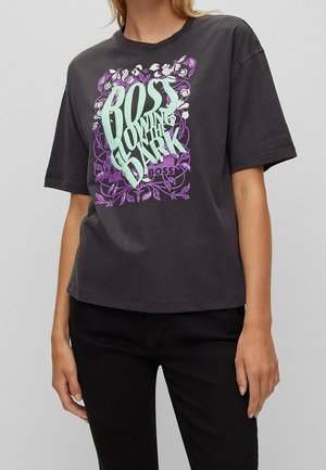 Mujer vistiendo una camiseta negra con diseño floral en púrpura y blanco y texto en verde claro "BOSS GLOWING IN THE DARK", combinada con pantalones negros.
