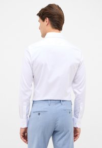 Witte long-sleeve shirt met textuurstof, klassieke kraag en knoopmanchetten, gecombineerd met lichtblauwe broek met zakken en subtiele details.